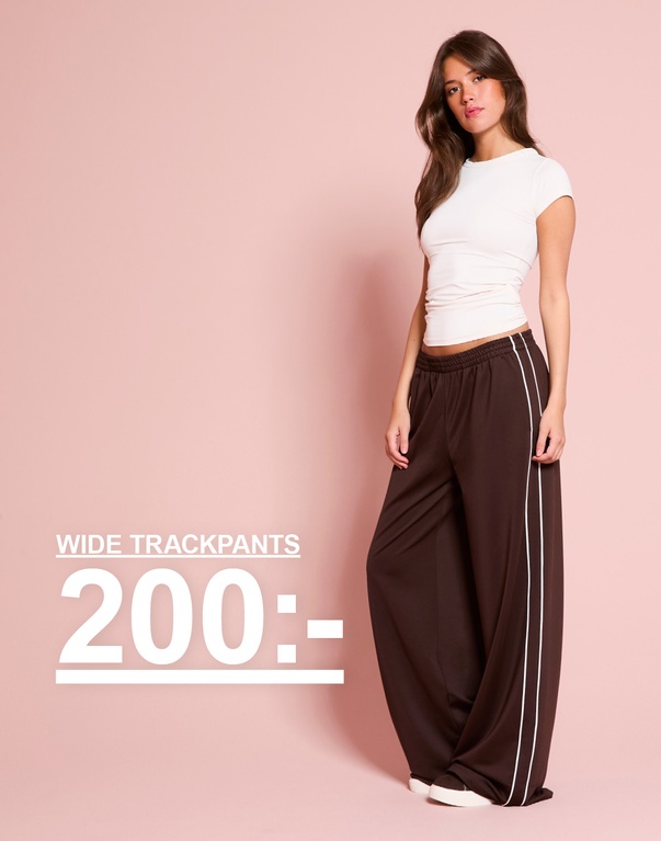 Tjej bär en vit t-shirt och bruna vida trackpants med vita ränder. WIDE TRACKPANTS 200:-.