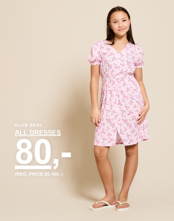 Barn i lyserød blomstret kjole og sandaler foran en lys baggrund. CLUB DEAL ALL DRESSES 80,- (REG. PRICE 80-160,-)