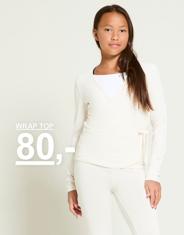 En pige iført et cremefarvet slå-om-top og matchende bukser står mod en lys baggrund. Tekst: Wrap top 80,-