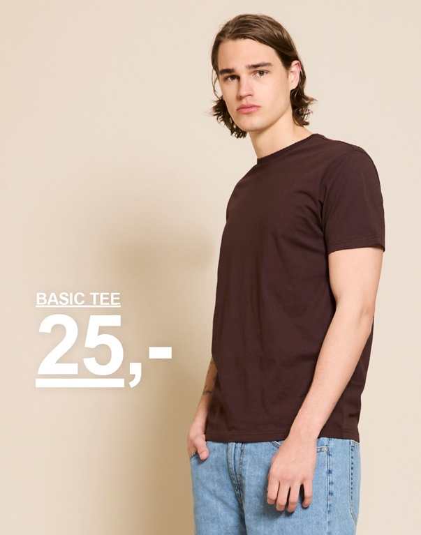 Model i mørkebrun basic t-shirt og lyse jeans foran beige baggrund. BASIC TEE 25,-