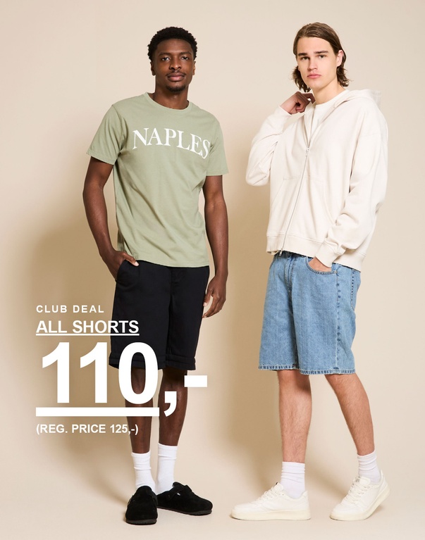 To mænd i shorts – den ene i grøn “NAPLES” T-shirt og mørke shorts, den anden i beige hoodie og lyse denimshorts foran en beige baggrund. CLUB DEAL. ALL SHORTS 110,- (REG. PRICE 125,-)