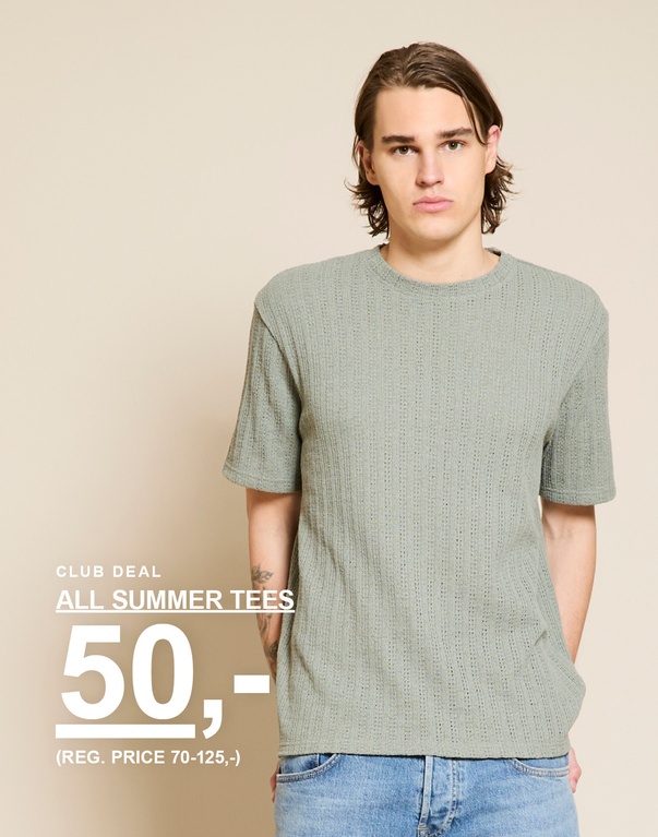 Mand i lysegrøn strikket T-shirt og lyseblå jeans mod en lys baggrund. CLUB DEAL. ALLE SOMMER-T-SHIRTS 50,- (FØRPRIS 70-125,-)