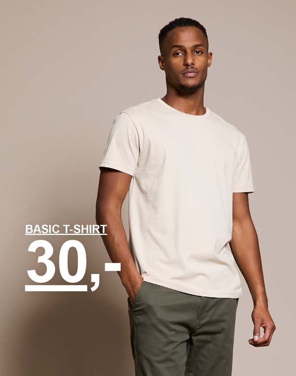 Mann i beige T-skjorte og grønne bukser står mot beige bakgrunn. Tekst på bildet viser: “Basic T-shirt 30,-”.