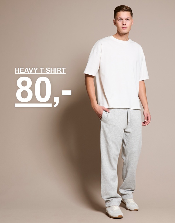 Mann i hvit T-skjorte og grå joggebukse står mot beige bakgrunn. Tekst på bildet viser: “Heavy T-shirt 80,-”.