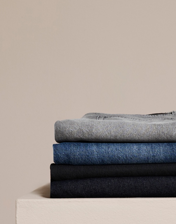 En stak foldede jeans i forskellige nuancer af blå og grå. Tekst: "DENIM" med information om et bredt udvalg af jeans til alle aldre og kropstyper med fokus på pasform, kvalitet og holdbarhed.