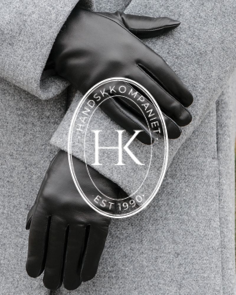 Svarta skinnhandskar som bärs till grå kappa, med rund logotyp handskkompaniet i bilden.
