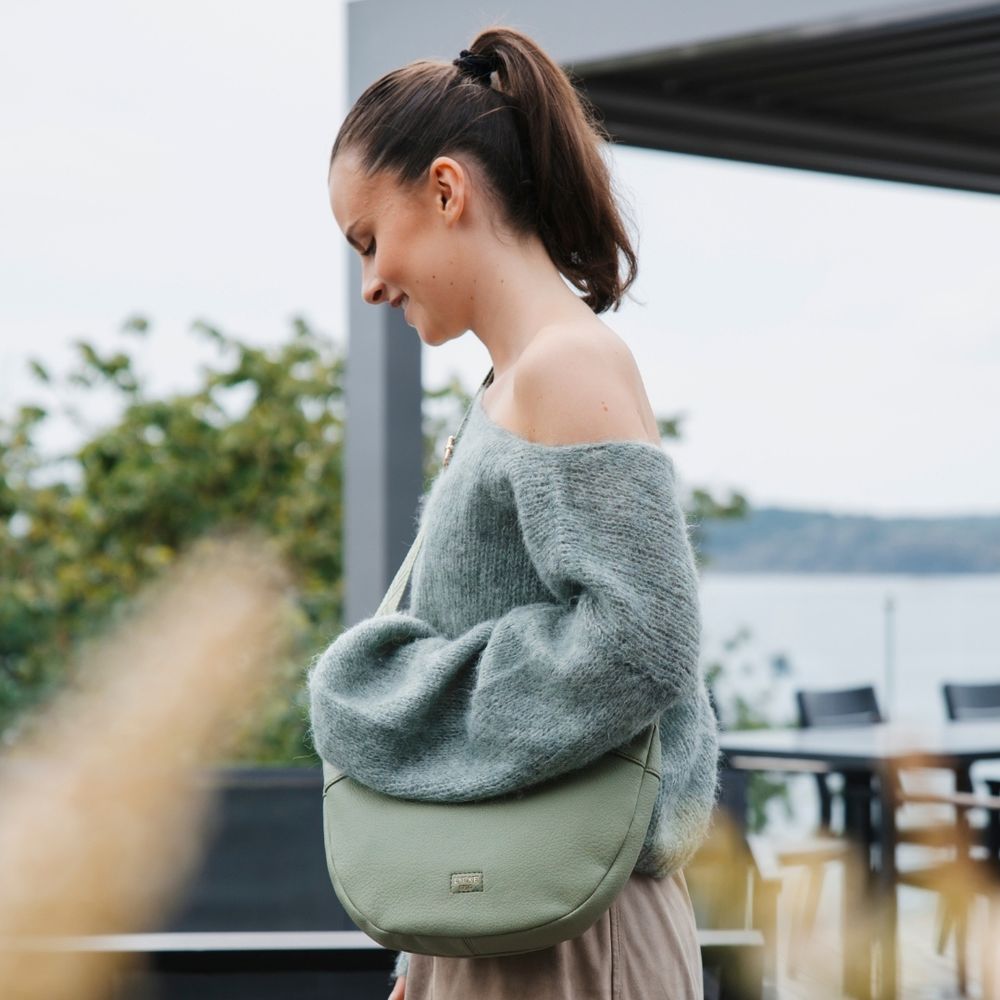 green crossover väska buren av kvinna i grå stickad tröja och beige kjol ute på en altan med havsutsikt