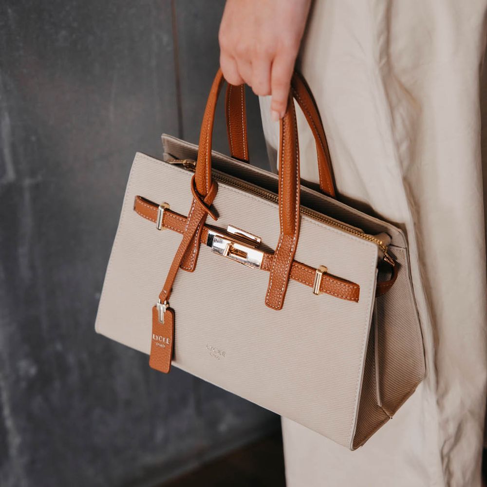 Beige handväska med bruna detaljer och dubbla handtag som hålls i handen. Lycke Oslo