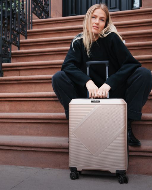 Dame sitter i en trapp i New York med en champagne farget koffert mellom bena
