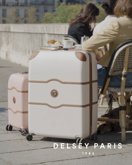 Lys beige Delsey Paris koffert med fire hjul plassert ved et kafébord utendørs