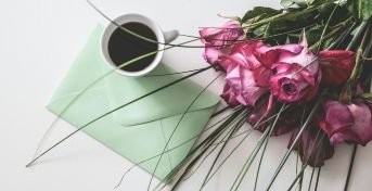 En rosa rosbukett liggandes på ett bord med ett grönt kuvert och en vit kaffekopp med kaffe i