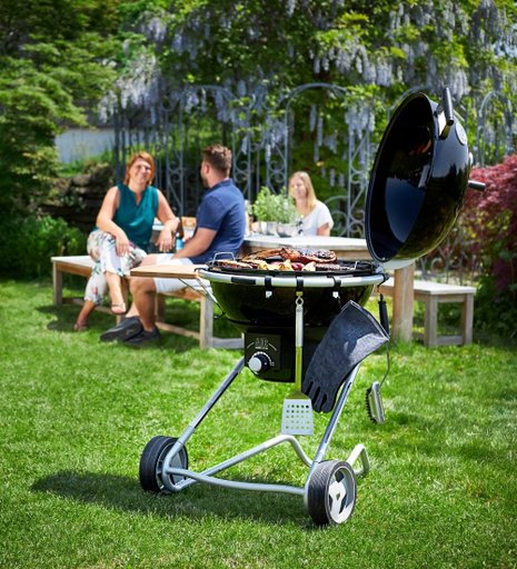 Ett svart kolgrill ståendes på en grön gräsmatta med människor i bakgrunden.