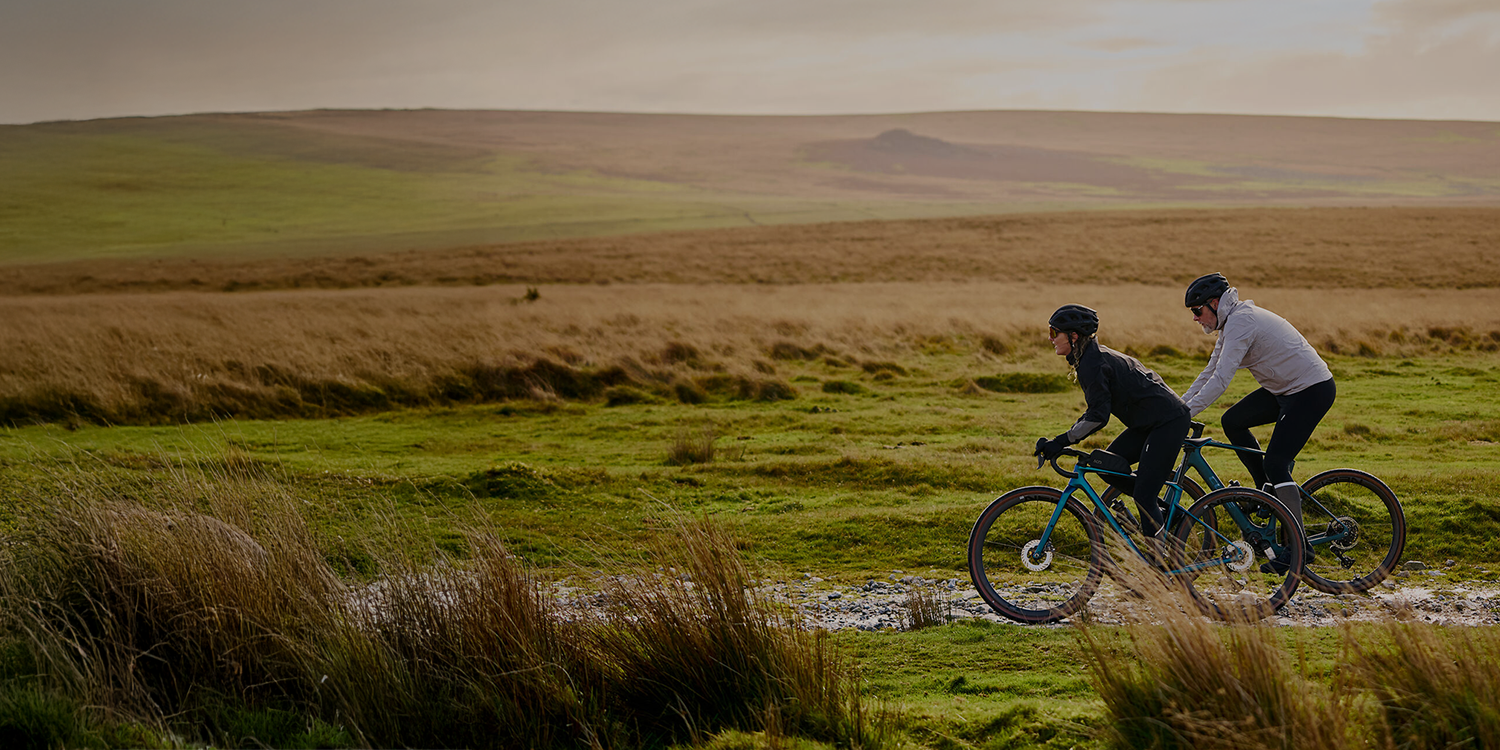 Pyöräilijä polkee ruskealla gravel-pyörällä leveää hiekkatietä, jonka reunoilla on metsää. Pyöräilijästä näkyy vain kädet ja jalat.