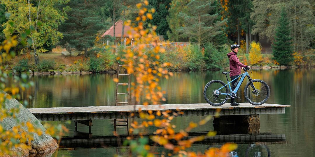 Pyöräilijä seisoo laiturilla järven rannalla vaaleansinisen Cube-sähkömaastopyörän kanssa