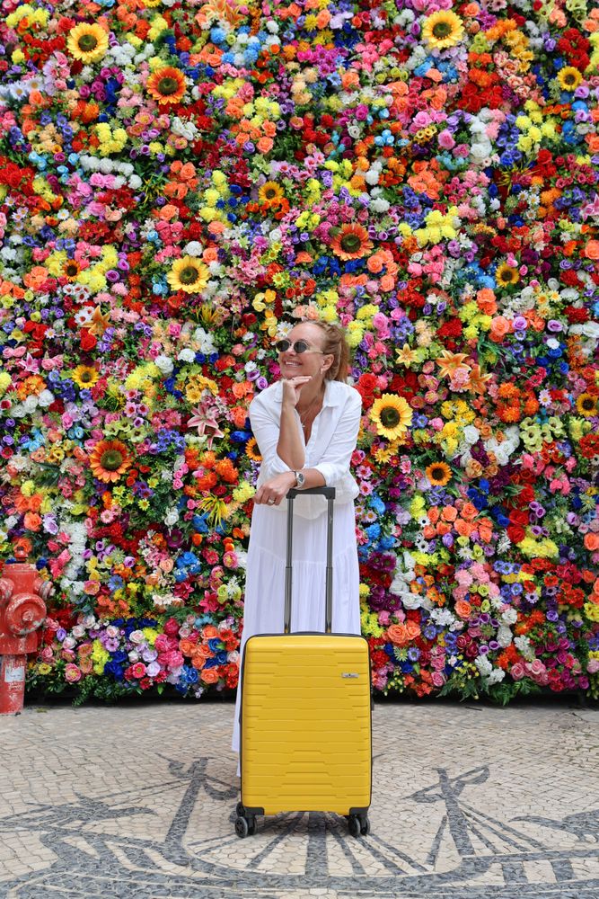 Dame i hvit kjole med gul koffert foran fargerik blomstrete vegg