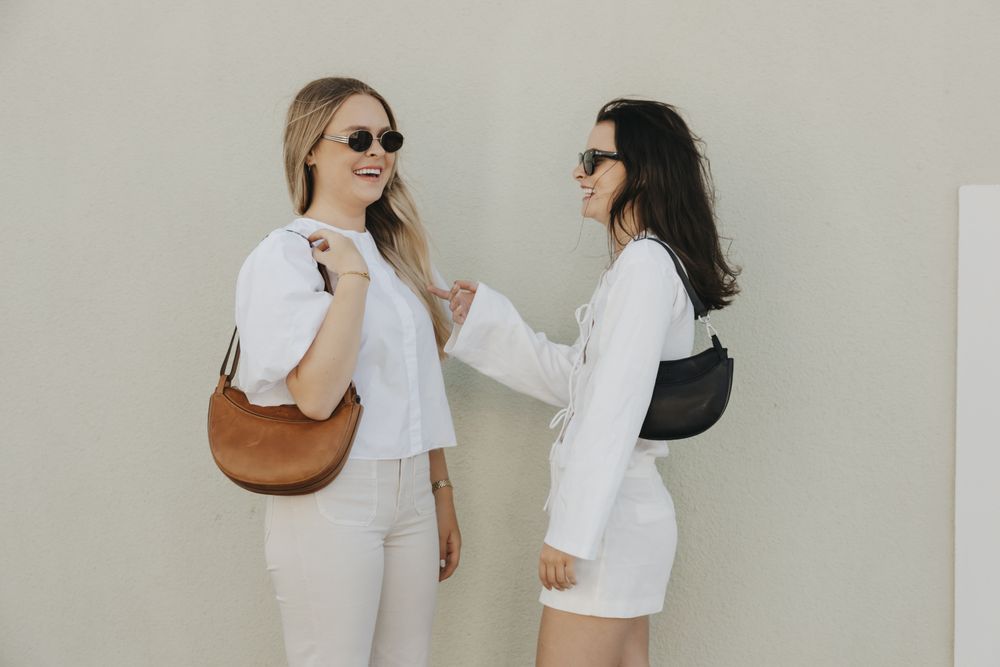 cognac skinnveske og svart skinnveske fra House of Nordic holdt av to damer kledd i vitt stående utendørs og ler