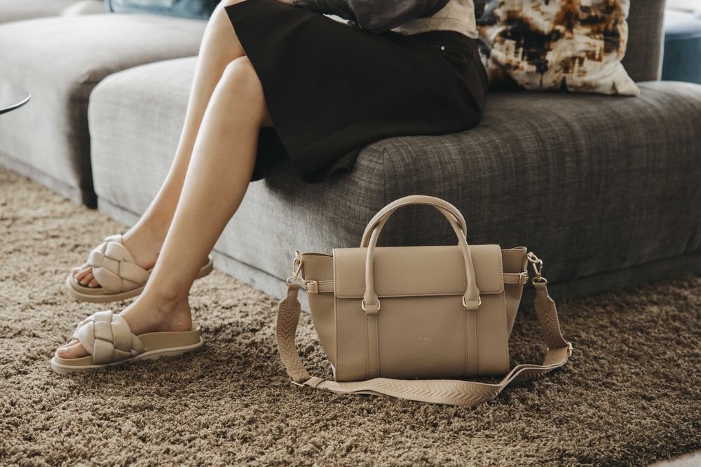 Dame sitter i en sofa med en beige håndveske på teppet ved siden av seg.