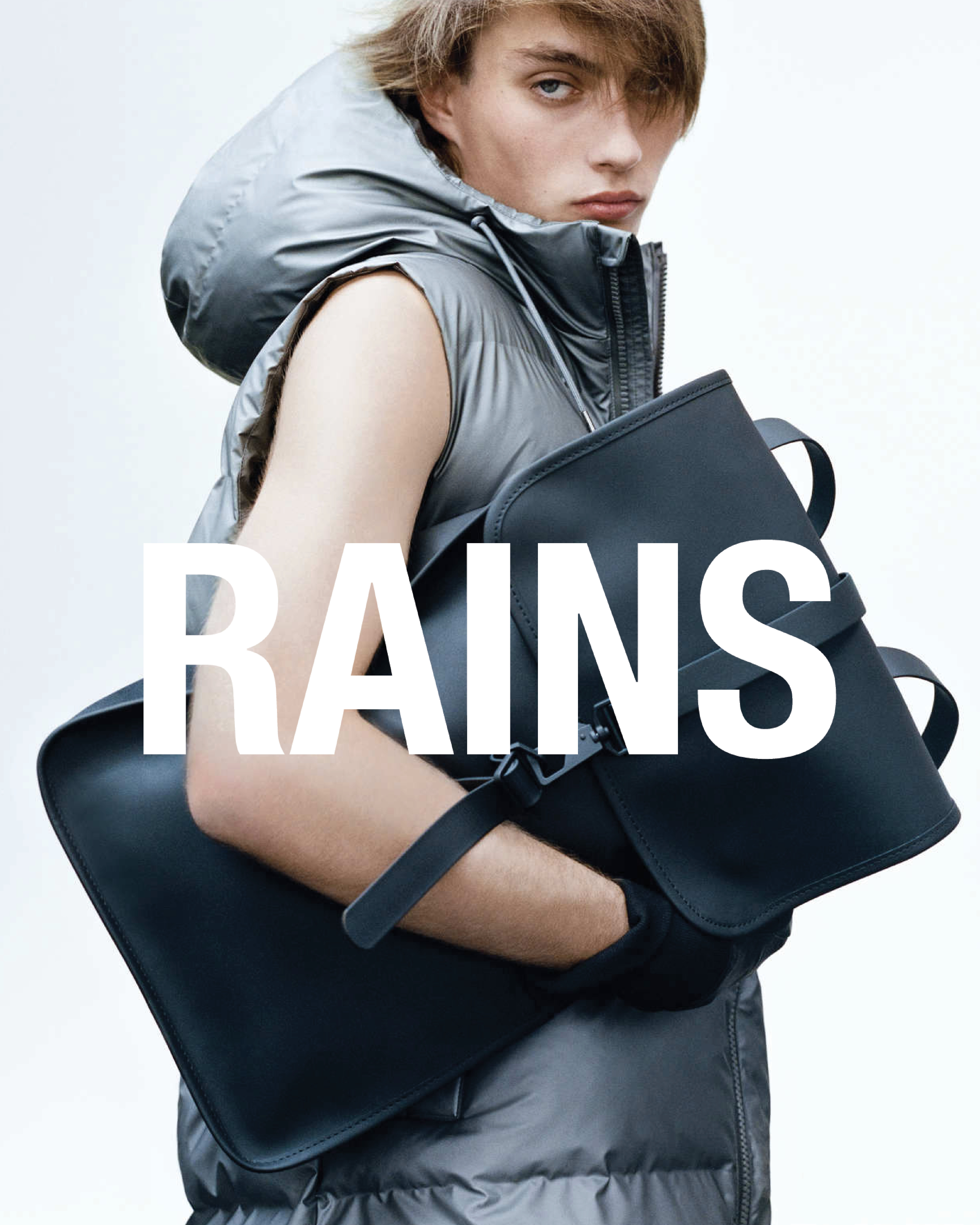 Person iført dunvest med ryggsekk under armen i urbant miljø, rains logo