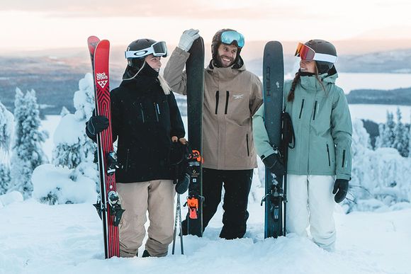 Tre personer står i snön med skidutrustning, skidjackor och skidbyxor som är ifrån Swedemount kollektionen St Anton