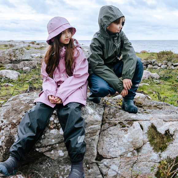 Tyttö ja poika istuvat kivellä sadevaatteet päällä