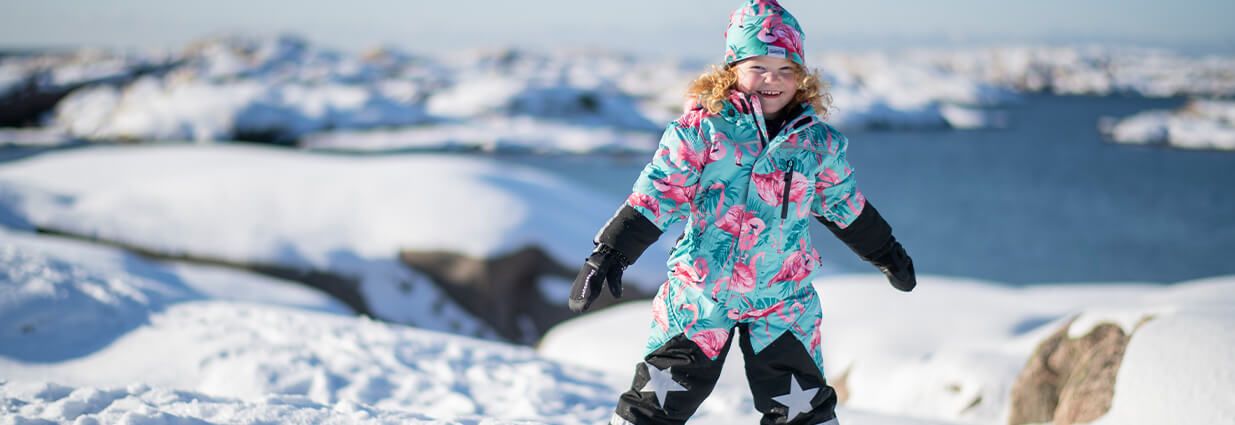 Ett barn/ junior som leker i snön med en Narvik overall ifrån Swedemount som är blå med rosa flamingos på