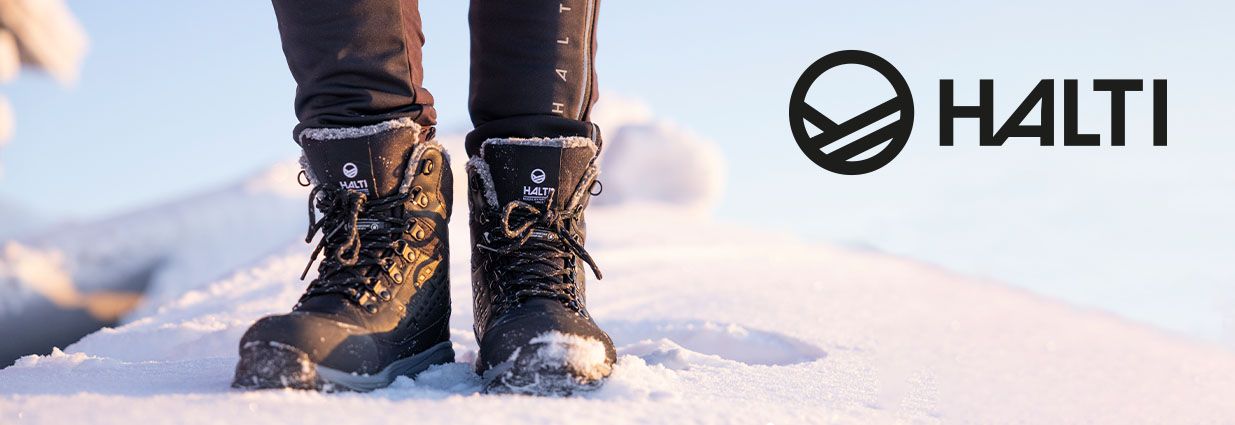 Henkilö seisoo lumessa Haltin talvikengät jalassa. Mustissa varsikengissä on nauhat ja Haltin logot. 