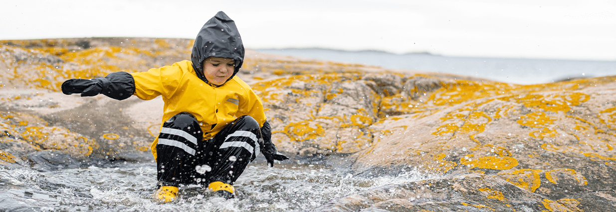 Et barn/junior hopper i sølepytter ved sjøen i gul og svart regnjakke fra Swedemount