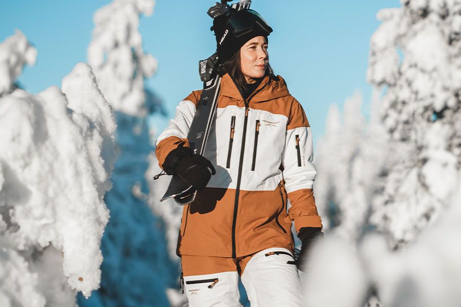 Nainen kantaa suksia lumisessa maisemassa ja hänellä on Swedemontin lasketteluhousut ja laskettelutakki päällä.