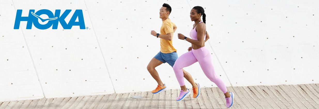 Mies ja nainen juoksevat valkoisen betoniseinän vieressä. Naisella on vaaleanpunaiset kengät ja vaatteet ja miehellä sinikeltaiset.