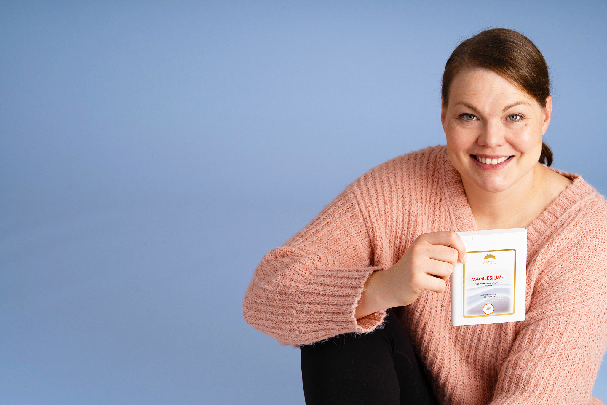 Smiling woman wearing a pink sweater holding Magnesium+ product